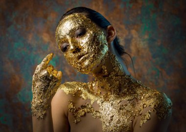 Yüzünde altın yapraktan yapılmış bir maske olan bir kız. Soyut bir arka planda esmer bir kadının kasvetli stüdyo portresi.
