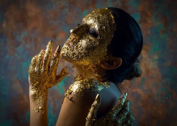 Girl with a mask on her face made of gold leaf. Gloomy studio portrait of a brunette on an abstract background.