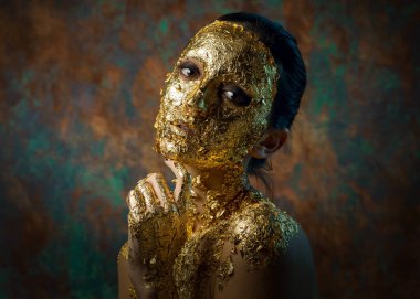 Yüzünde altın yapraktan yapılmış bir maske olan bir kız. Soyut bir arka planda esmer bir kadının kasvetli stüdyo portresi.