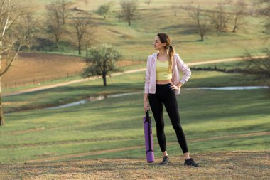 Spor giyimli genç, zayıf, atletik bir kız bir takım egzersizler yapıyor. Yeşil bahar otlak tepelerinin arka planında sağlık ve sağlıklı yaşam tarzı.