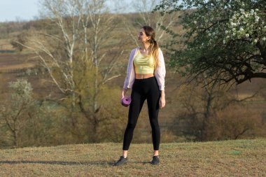 Spor giyimli genç, zayıf, atletik bir kız bir takım egzersizler yapıyor. Yeşil bahar otlak tepelerinin arka planında sağlık ve sağlıklı yaşam tarzı.