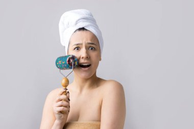 Kafasında havlu olan esmer, gri arka planda izole edilmiş yüz masajı için silindir kullanıyor.