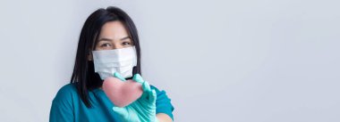Koruyucu tıbbi maske takan esmer genç bir kız pembe bir kalp taşıyor, hayatı simgeliyor. Coronavirus konsepti.