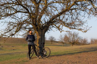 Şortlu ve formalı bisikletçi, modern karbon kuyruklu motorsikletli. Uçurumun kenarında, yeşil bahar ormanlarının arka planında duran bir hava süspansiyon çatalı var.