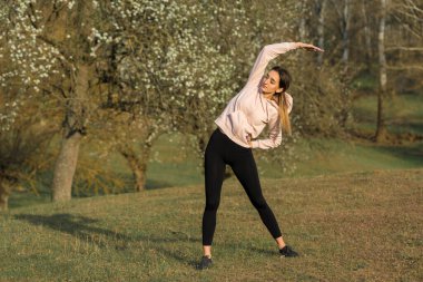 Spor giyimli genç, zayıf, atletik bir kız bir takım egzersizler yapıyor. Yeşil bahar otlak tepelerinin arka planında sağlık ve sağlıklı yaşam tarzı.