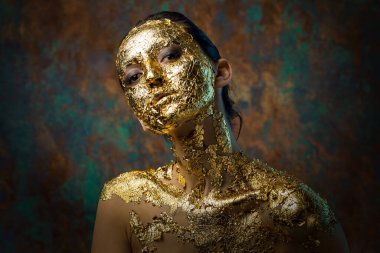 Yüzünde altın yapraktan yapılmış bir maske olan bir kız. Soyut bir arka planda esmer bir kadının kasvetli stüdyo portresi.