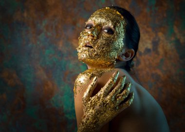 Yüzünde altın yapraktan yapılmış bir maske olan bir kız. Soyut bir arka planda esmer bir kadının kasvetli stüdyo portresi.