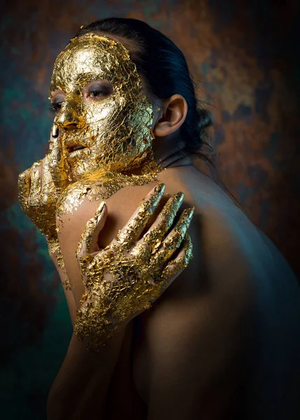 Girl with a mask on her face made of gold leaf. Gloomy studio portrait of a brunette on an abstract background.