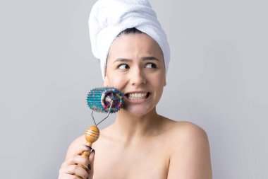 Kafasında havlu olan esmer, gri arka planda izole edilmiş yüz masajı için silindir kullanıyor.