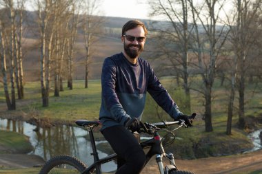 Şortlu ve formalı bisikletçi, modern karbon kuyruklu motorsikletli. Uçurumun kenarında, yeşil bahar ormanlarının arka planında duran bir hava süspansiyon çatalı var.