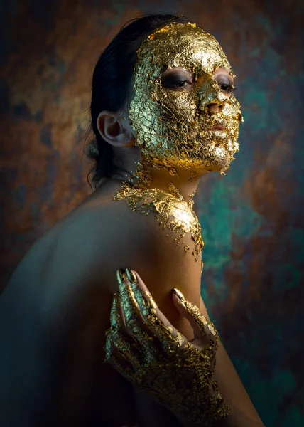 Girl with a mask on her face made of gold leaf. Gloomy studio portrait of a brunette on an abstract background.