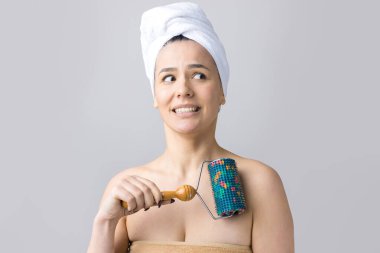 Kafasında havlu olan esmer, gri arka planda izole edilmiş yüz masajı için silindir kullanıyor.
