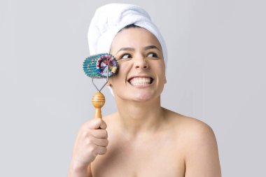 Kafasında havlu olan esmer, gri arka planda izole edilmiş yüz masajı için silindir kullanıyor.