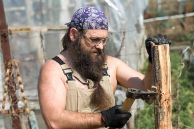 Bandanalı orta yaşlı sakallı bir adam baltayla kütük kesiyor. Tulumlu gaddar sıcak bir günde zor işi yapar..