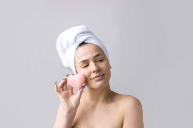 Kafasında beyaz havluyla güzel bir kadın portresi ve pembe bir kalbe bakan bir vücut için sünger. Cilt temizleme eko organik kozmetik rahatlama konsepti.