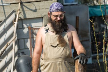 Bandanalı orta yaşlı sakallı bir adam baltayla kütük kesiyor. Tulumlu gaddar sıcak bir günde zor işi yapar..