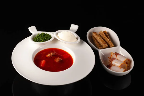 Ukrainian borsch with bacon and crispbread in a white plate on a black background