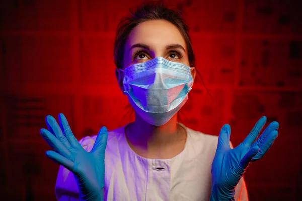 Medical worker in a medical mask and rubber gloves. medic girl in a dark room on a red background. medical practitioner in despair during the coronavria pandemic.