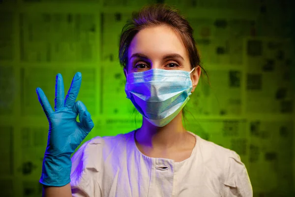 Maskeli ve eldivenli mutlu doktor kız her şeyin yolunda olduğunu gösteriyor. Tıbbi maske takan ve yeşil arka planda lastik eldiven takan bir tıp çalışanı. Koronavirüs salgınına karşı zafer