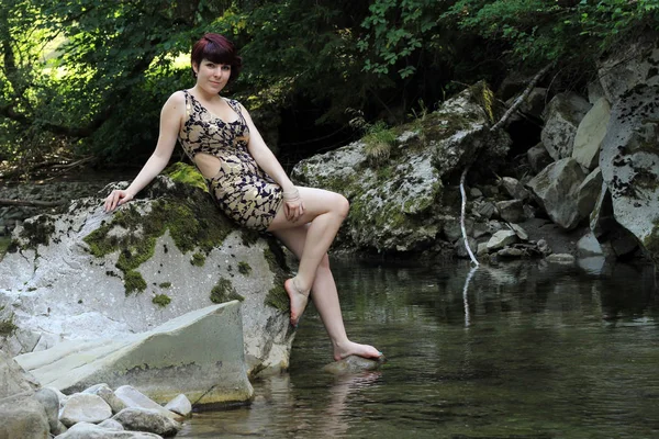 一个穿着迷你连衣裙的漂亮年轻女子坐在小溪边的石头上 — 图库照片