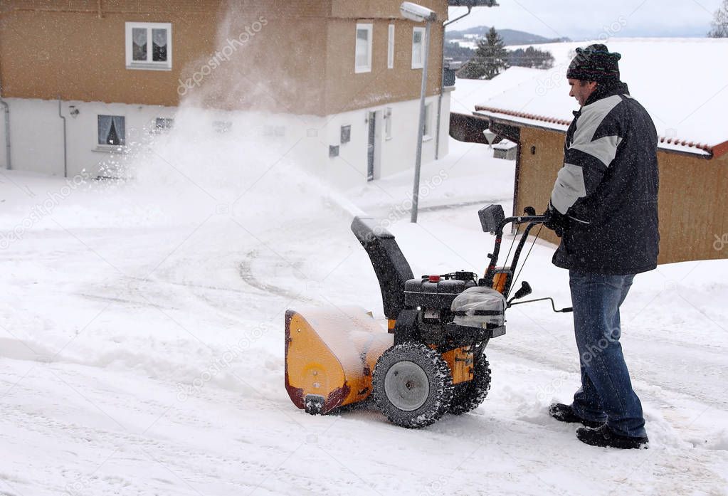 A man clears the road and the sidewalk with a snow blower