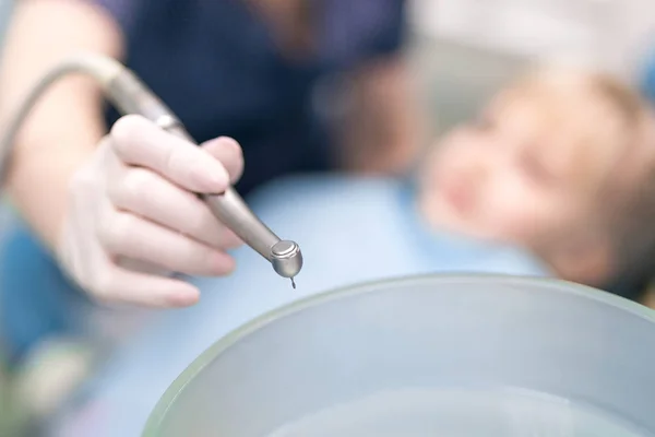 Close-up van artsen hand met tandheelkundige boor machine. Wazig kind in stoel stomatologie kliniek op achtergrond. Kinderen bezoeken stomatologie angst of fobie concept. Tand gezondheidszorg concept — Stockfoto