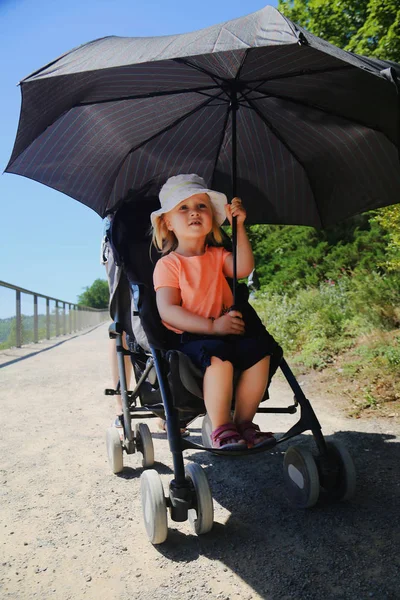 Mała dziewczynka siedzi w wózek. Dziecko chronione przed słońcem przez duży parasol. Zapobiegania udar cieplny dziecko na słoneczny dzień w lecie — Zdjęcie stockowe