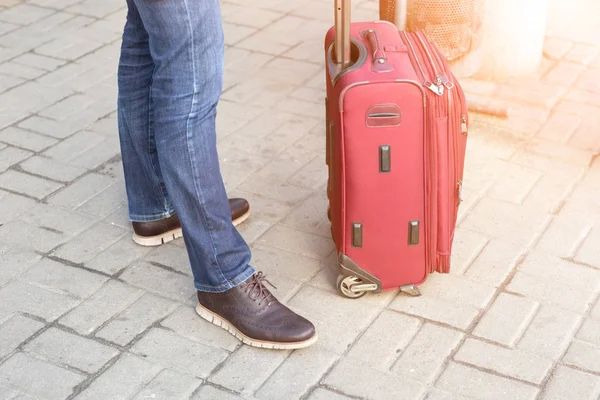 Мужчина в повседневной одежде стоит с красным чемоданом на станции. Молодой современный путешественник ждет удобного путешествия. Человек с багажом — стоковое фото
