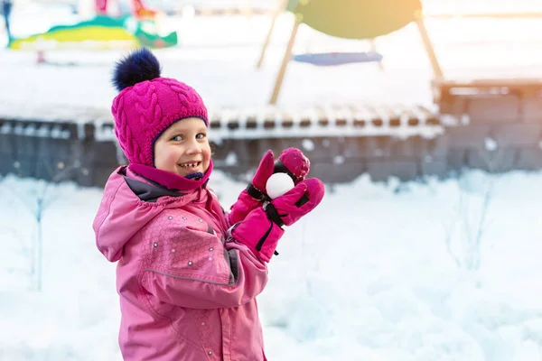 Linda adorable niña caucásica retrato de invierno sosteniendo bola de nieve en las manos listo para la lucha de nieve en el patio al aire libre. Divertido niño juguetón durante las nevadas en la estación fría al aire libre. Feliz infancia. —  Fotos de Stock