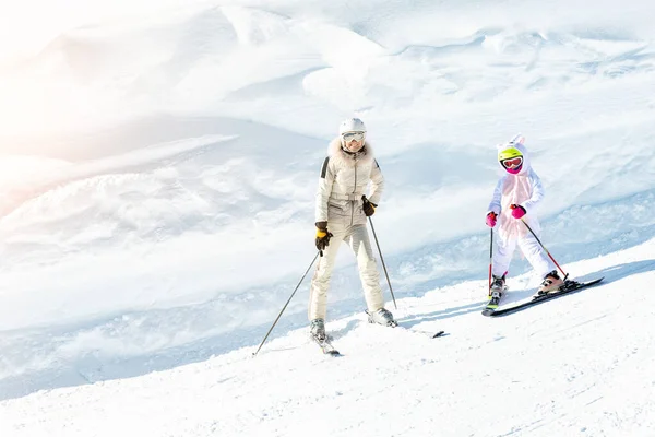 Young adult beautiful sporty mother having fun skiing with kid daughter on mountain alpine ski winter resort. Slim mom in luxury fashion skier suit and child enjoy outdoor recreation activities — Stock Photo, Image