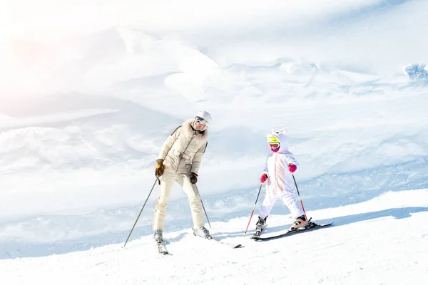 Young adult beautiful sporty mother having fun skiing with kid daughter on mountain alpine ski winter resort. Slim mom in luxury fashion skier suit and child enjoy outdoor recreation activities — Stock Photo, Image