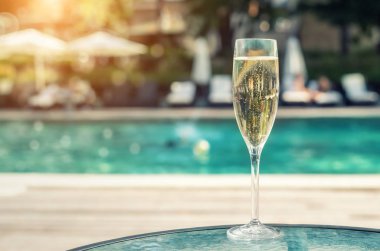 Tatil sırasında lüks tatil otelinde havuz kenarına karşı yakın çekim beyaz şampanya veya prosecco cam. Açık mavi havuz arka plan ile yükselen kabarcıklar ile köpüklü şarap. Ferahlatıcı alkol içeceği