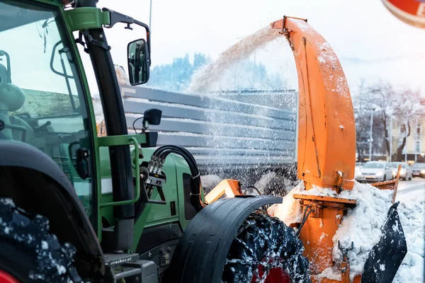 Gran tractor con cadenas en la rueda que sopla nieve de la calle de la ciudad en el cuerpo del camión de volteo con el ventilador de nieve. Limpieza de calles y remoción de nieve después de nevadas. Servicios municipales remoción de nieve en temporada de invierno — Foto de Stock