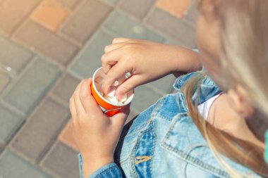 Kapalı kafkasyalı, kot ceketli küçük sarışın kız dışarıda yürürken kağıt bardaktan tatlı küçük şekerlemeler yiyor. Sağlıksız abur cubur ve çocuk obezitesi