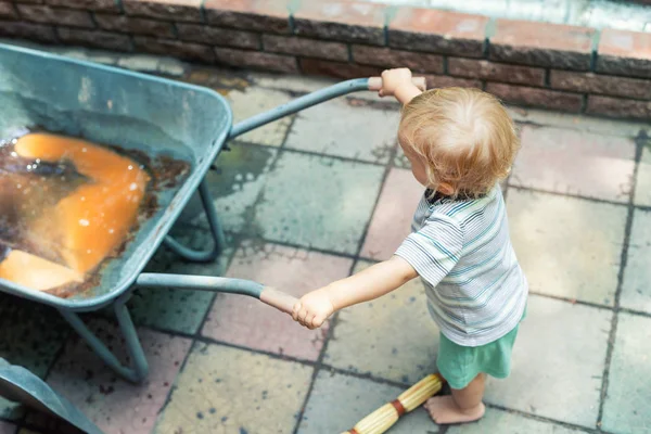 Bonito adorável caucasiano criança menino brincando com grande velho carrinho de mão no quintal no jardim ao ar livre. Criança pequeno ajudante em t-short e shorts se divertindo empurrando carrinho de mão e jardinagem no campo — Fotografia de Stock