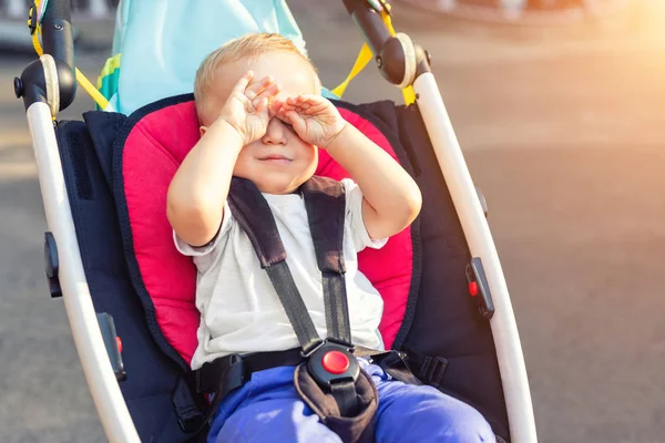 Pouco bonito adorável criança garoto esfregando os olhos com as mãos sentadas em carrinho no dia brilhante de verão ao ar livre. A criança cansada quer dormir durante o passeio na carruagem do lado de fora. Bebê tentando dormir — Fotografia de Stock