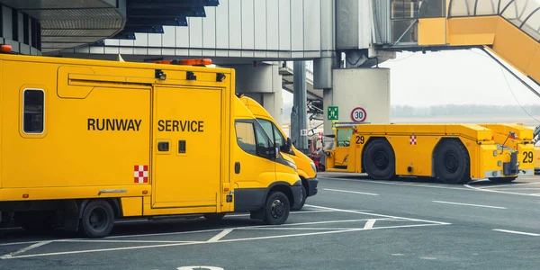 Runway service voertuigen staan op asfalt in de buurt van luchthaven terminal gebouw. Speciale onderhoudsploeg vrachtwagen bestelwagen en trekker geparkeerd op gemarkeerde plaats parkeerplaats. Nood- en ondersteuningsapparatuur op het vliegveld — Stockfoto