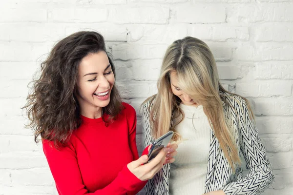 İki yetişkin beyaz kız arkadaş arka plandaki beyaz tuğla duvara karşı eğleniyor ve birlikte vakit geçiriyorlar. Evde sosyal ağlar için selfie çekerken gülümseyen iki arkadaş.