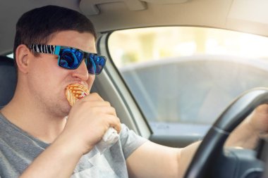 Güneşli bir günde araba sürerken yetişkin bir erkek beyaz adam sosisli fast food yiyor. Erkekler direksiyon başında yemek yiyorlar. Araçtaki dikkati dağılmış adam. Kötü bir alışkanlık. Abur cubur.