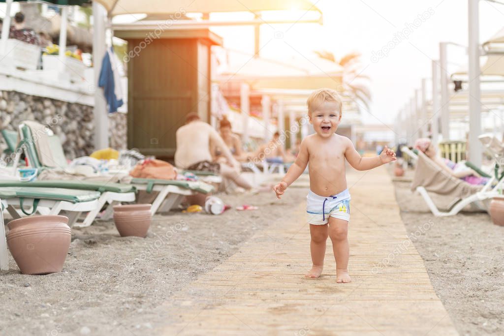 Cute caucasian toodler boy walking alone on sandy beach between chaise-lounge. Adorable happy child having fun playing at seaside shore during vacation trip