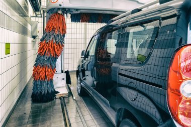 Robot otomatik tünel oto yıkama makinesiyle siyah, modern minibüs araba temizliği. Benzin istasyonunda köpükle yıkanan gri ve kırmızı fırçalar. Araba yıkama işi.
