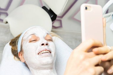 Genç yetişkin bir kadın güzellik salonunda yüz maskesi takmış bir şekilde yatakta uzanıyor ve sosyal ağlar için telefon özçekimi yapıyor. Cilt bakımı ürün değerlendirmesi ile çevrimiçi vlog akışı yapan nüfuz edici