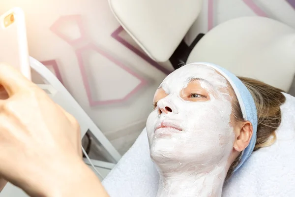 Mujer joven adulta acostada en la cama en el salón de spa de belleza con máscara cosmética facial aplicada haciendo foto selfie teléfono para las redes sociales. Influencer haciendo flujo vlog en línea con revisión del producto de cuidado de la piel —  Fotos de Stock