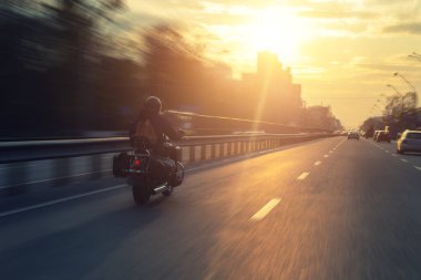 Şehir caddesinde siyah motosiklet kullanan motorcu silueti ve arka planda günbatımı dramatik gökyüzü. Yalnız motorcuların kurutma döngüsü. Seyahat macerası ve seyahat konsepti. Yaşam biçimi