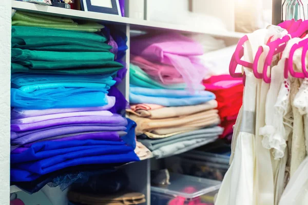 Rack with many beautiful holiday dresses for girls on hangers and multicolored fabric stack at children fashion sewing studio and showroom indoor. Female dress rental salon maker photo session event — Stock Photo, Image