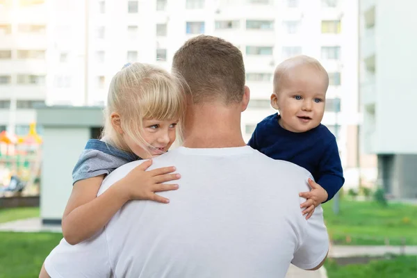 Widok z tyłu ojca trzymającego dwójkę uroczych słodkich rodzeństwa na rękach podczas spaceru na świeżym powietrzu. Mały syn i córka cieszą się i bawią przytulając tatusia w letni dzień. Koncepcja rodziny i opieki nad dziećmi — Zdjęcie stockowe