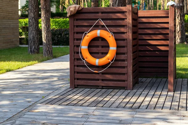 Ξύλινα αποδυτήρια με πορτοκαλί ναυαγοσωστικό σε χώρο αναψυχής κοντά σε λίμνη, πισίνα ή λίμνη στο δασικό οικολογικό θέρετρο. Καμπίνα σε ξύλινο δάπεδο και γκαζόν κοντά στην πισίνα σε πευκοδάσος — Φωτογραφία Αρχείου