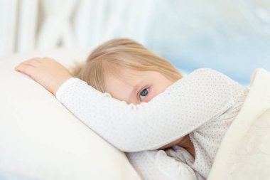 Beyaz yastıkla yatakta uzanan sevimli, sevimli, beyaz, beyaz, beyaz, küçük, sarışın bir kız. Elle yüzünü örten üzgün küçük bir çocuğun portresi. Mutsuz çocuk sabah erken kalkmaktan memnun değil..