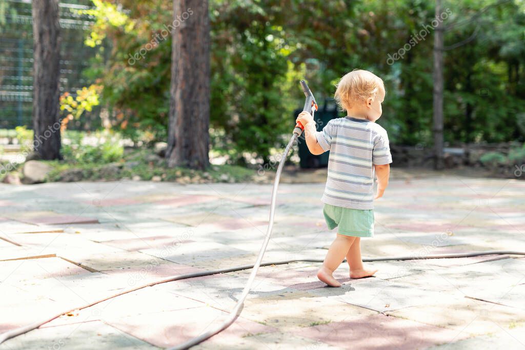 Cute adorable caucasian blond barefeet boy walking at home backyard holding hose pipe for watering garden. Child little helper playing gardening at summer outdoors.