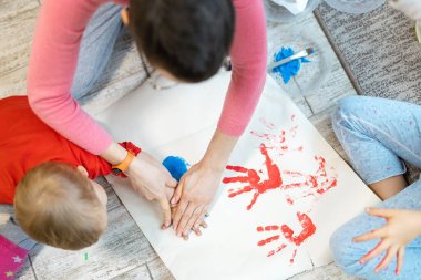 Sevimli, beyaz, sarışın küçük kardeşler anneleriyle evde fırça ve avuç içi boyama yaparken eğleniyorlar. Neşeli mutlu çocuklar resim çizerek gülümsüyor..